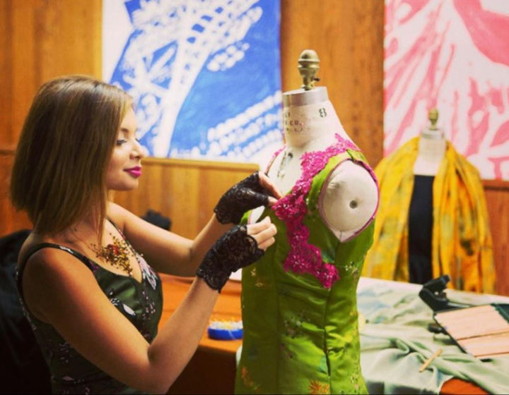 Dona pinning a piece of pink lace on a green satin dress on a dressform in fashion school