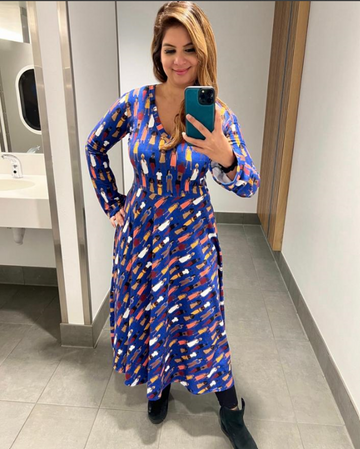 Dona doing a mirror selfie wearing the Manila dress. Long-sleeved, Midi-length. Bright cobalt blue background with a foreground of sketches of women standing with power.
